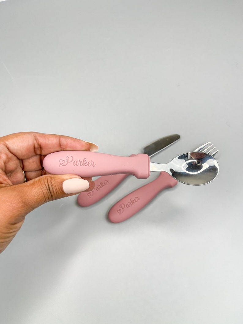 May include: A set of three pink baby utensils with a silicone handle. The utensils are a fork, spoon, and knife. The handle has the name "Parker" engraved on it.