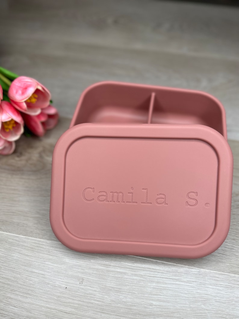 May include: A pink silicone lunch box with a divided interior and a personalized inscription that reads "Camila S."