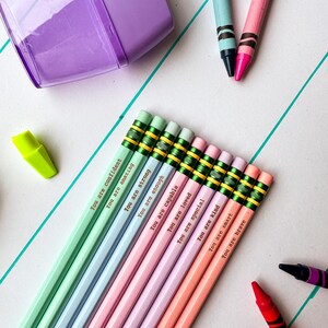Puede incluir: Una colección de lápices de colores pastel con frases motivacionales como "You are confident" y "You are brave." Los lápices están dispuestos con un sacapuntas morado, crayones y un resaltador sobre una superficie blanca con líneas turquesas.