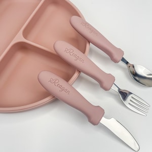 May include: A pink silicone divided plate with a set of three pink handled utensils. The utensils are a knife, fork, and spoon. The handles of the utensils are engraved with the name "Reagan".