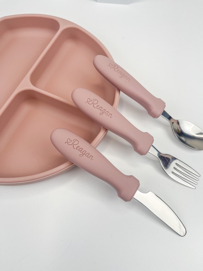 May include: A pink silicone divided plate with a set of three pink handled utensils. The utensils are a knife, fork, and spoon. The handles of the utensils are engraved with the name "Reagan".