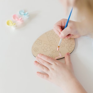 Puede incluir: Un niño pinta un huevo de Pascua de madera con pintura rosa. El huevo está decorado con flores y la palabra "Otoño" está escrita en él.