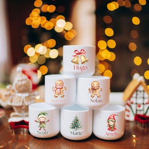 May include: White ceramic mugs with Christmas-themed illustrations and names. Designs include gingerbread people, an elf, a Christmas tree, and bells. Names on the mugs are Hugo, Sammy, Nolan, Noah, Marie, and Amelia. Festive holiday decor.