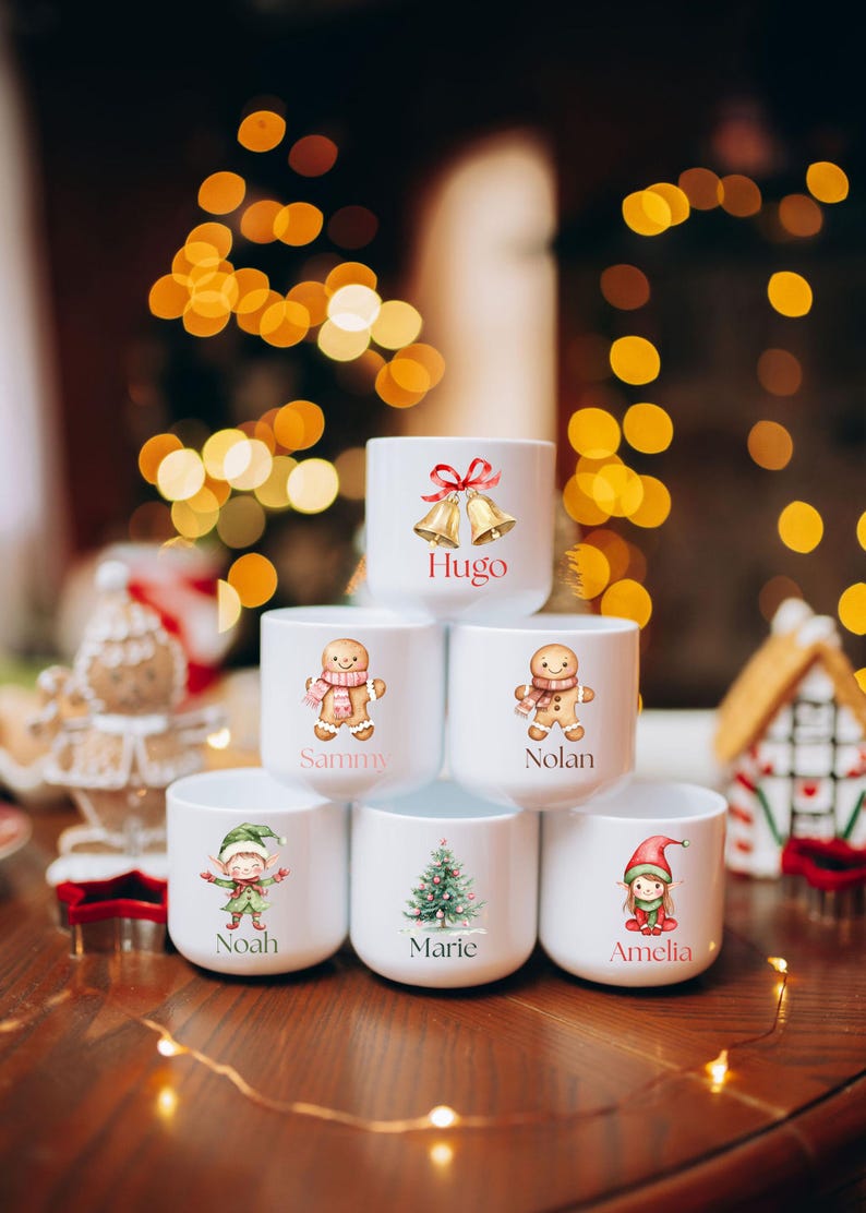 May include: White ceramic mugs with Christmas-themed illustrations and names. Designs include gingerbread people, an elf, a Christmas tree, and bells. Names on the mugs are Hugo, Sammy, Nolan, Noah, Marie, and Amelia. Festive holiday decor.