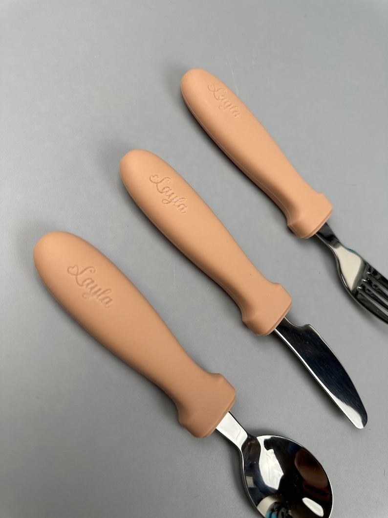 May include: A set of three personalized cutlery with beige silicone handles. The handles are engraved with the name "Layla". The set includes a fork, knife, and spoon.