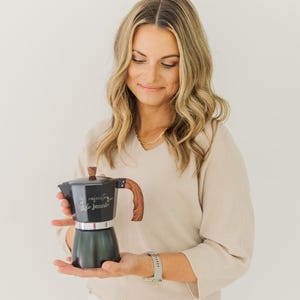 May include: A black and silver stovetop espresso maker with a wooden handle and lid. The espresso maker has the text "cafecito by Jennie" on the side. The handle and lid are made of wood. The espresso maker is held in front of a white background.