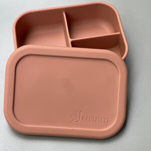 May include: A light pink silicone lunch box with three compartments and a lid. The lid has the word "Journey" embossed on it.
