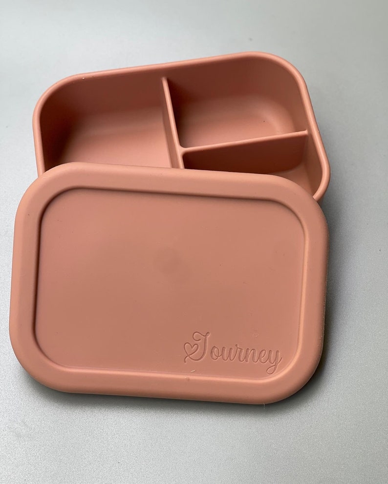 May include: A light pink silicone lunch box with three compartments and a lid. The lid has the word "Journey" embossed on it.