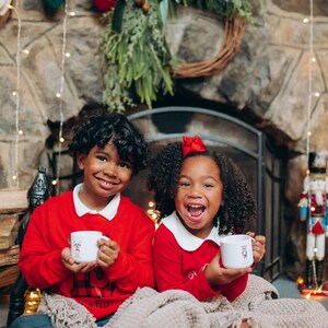 May include: Two children in red sweaters with white collars and mugs, smiling in front of a fireplace decorated for Christmas. A beige blanket covers their legs. A wreath and stockings hang above the fireplace. A nutcracker is visible.