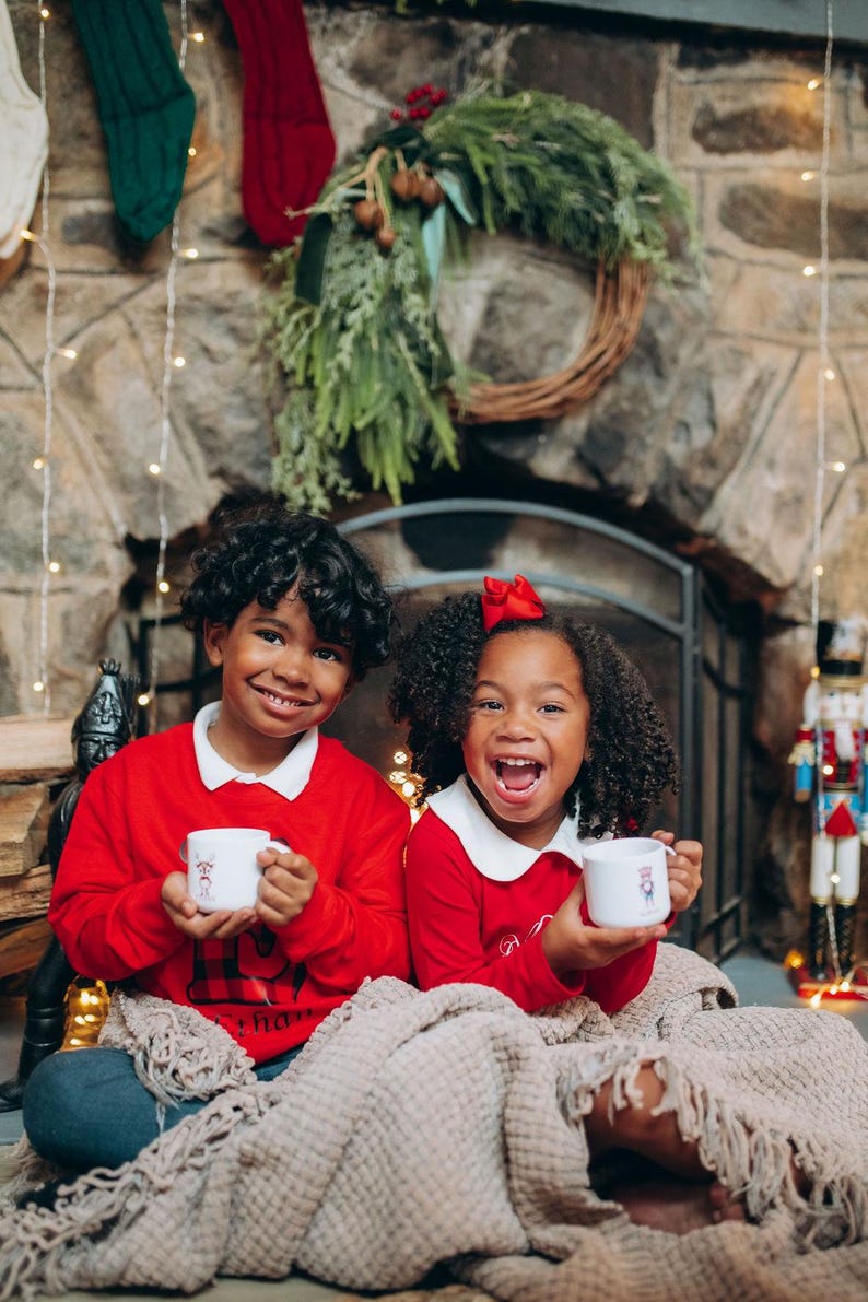 May include: Two children in red sweaters with white collars and mugs, smiling in front of a fireplace decorated for Christmas. A beige blanket covers their legs. A wreath and stockings hang above the fireplace. A nutcracker is visible.