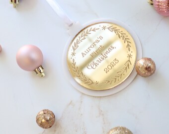 May include: Gold Christmas ornament with a white background. The ornament is round and has a white ribbon attached. The ornament is engraved with the text "Aurora's First Christmas, 2023".