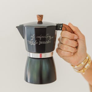 May include: Black stovetop espresso maker with a wooden handle and top. The text "el cafecito de Jennie" is etched on the side. The espresso maker is held by a person wearing gold bracelets.