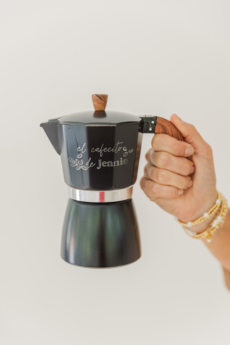 May include: Black stovetop espresso maker with a wooden handle and top. The text "el cafecito de Jennie" is etched on the side. The espresso maker is held by a person wearing gold bracelets.