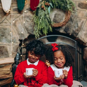 May include: Two children in red sweaters with white collars hold mugs with a holiday design. They are sitting in front of a stone fireplace decorated with a wreath, stockings, and lights. A cozy blanket is draped over their laps.