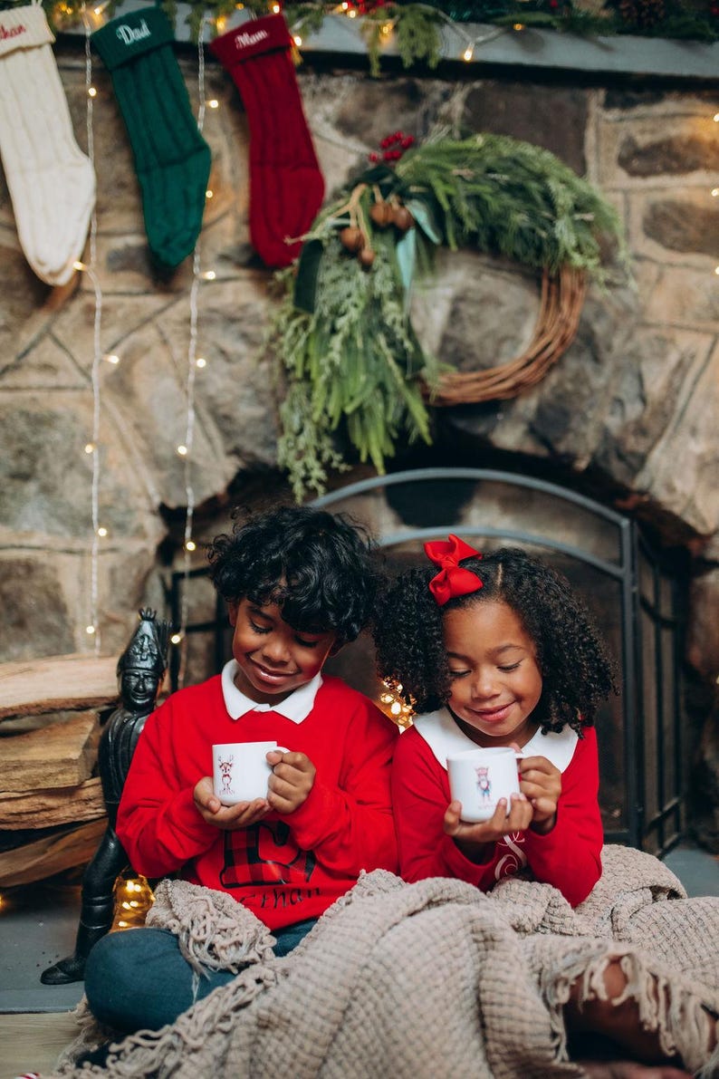 May include: Two children in red sweaters with white collars hold mugs with a holiday design. They are sitting in front of a stone fireplace decorated with a wreath, stockings, and lights. A cozy blanket is draped over their laps.