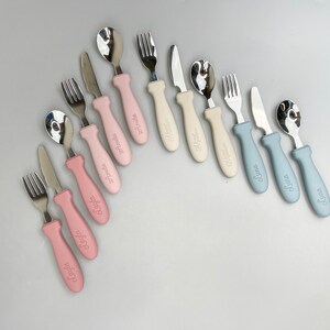 May include: A set of nine personalized baby cutlery sets with pink, blue, and beige handles. Each set includes a spoon, fork, and knife with a name engraved on the handle.