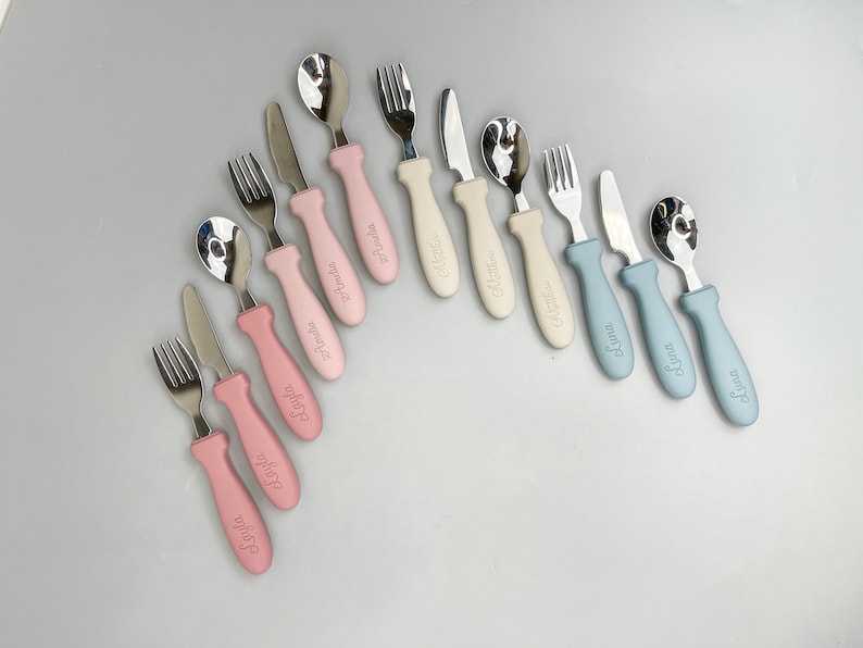 May include: A set of nine personalized baby cutlery sets with pink, blue, and beige handles. Each set includes a spoon, fork, and knife with a name engraved on the handle.