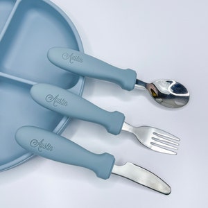 May include: A blue silicone baby plate with three compartments and a matching blue silicone spoon, fork, and knife with the name "Austin" engraved on the handles.