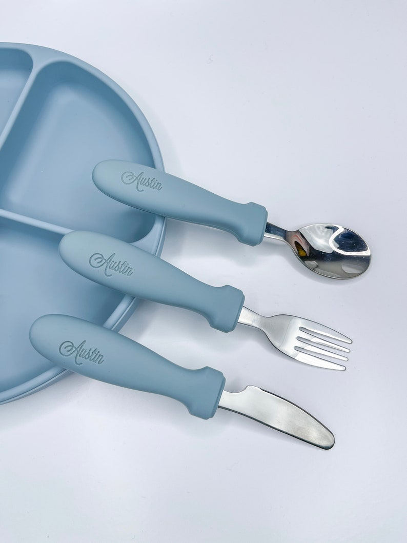 May include: A blue silicone baby plate with three compartments and a matching blue silicone spoon, fork, and knife with the name "Austin" engraved on the handles.