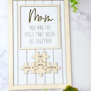 Puede incluir: Un marco de madera con fondo blanco y el texto "Mom You are the piece that holds us together". Debajo del texto hay cuatro piezas de rompecabezas de madera que dicen "Dad", "Mom", "Hugo E." y "Sophia".