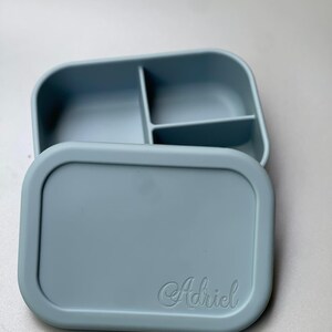 May include: A light blue silicone lunch box with three compartments and a lid. The lid has the name "Adriel" embossed on it.