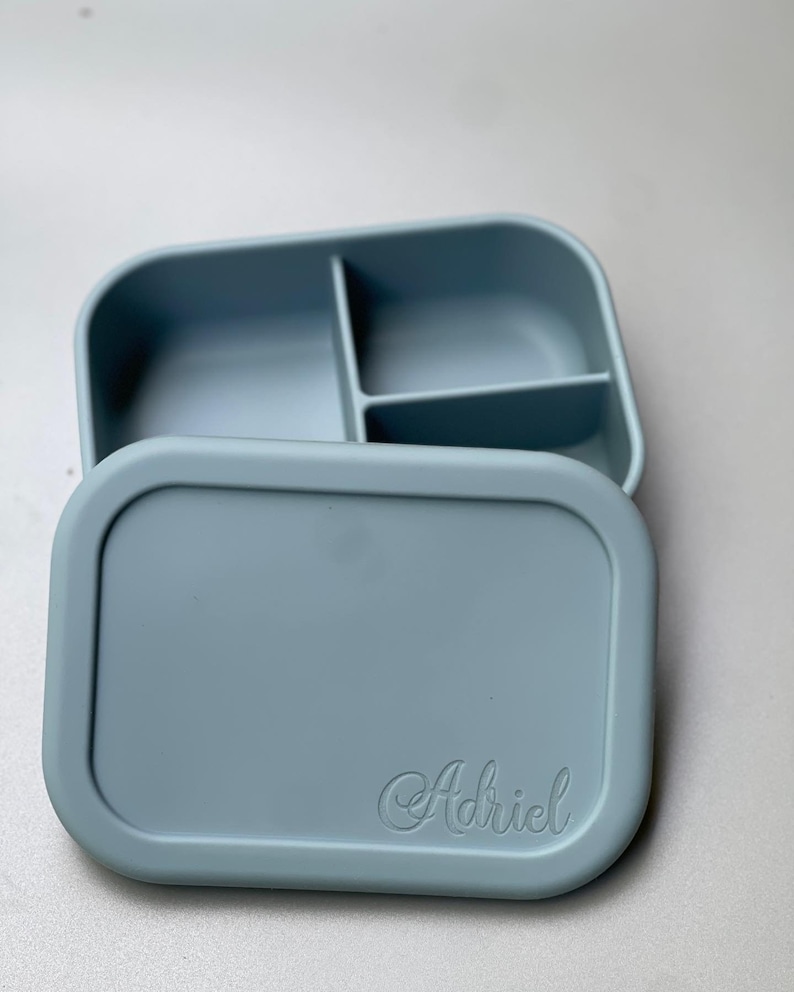 May include: A light blue silicone lunch box with three compartments and a lid. The lid has the name "Adriel" embossed on it.