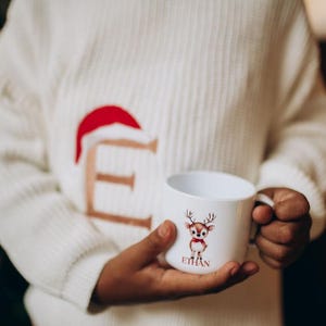 May include: A white ceramic mug featuring a cartoon reindeer with a red bow tie and the name "Ethan" printed below. The mug is held by a person wearing a white sweater with a red Santa hat design.