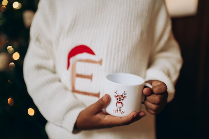 May include: A white ceramic mug featuring a cartoon reindeer with a red bow tie and the name "Ethan" printed below. The mug is held by a person wearing a white sweater with a red Santa hat design.
