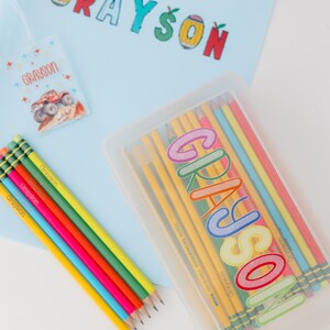 May include: A light blue binder, pencils, and a pencil case with the name "Grayson" in colorful letters. A matching tag with the name "Grayson" and a monster truck graphic is also present. The pencils are yellow, with the name "Grayson" printed on them.