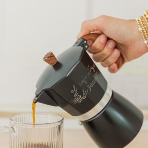 May include: A black stovetop espresso maker with a wooden handle and lid is pouring coffee into a clear glass mug. The espresso maker has the text "el cafecito de Jennie" on the side.