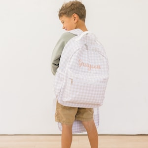 May include: A light-colored backpack with a gingham pattern and the name "Grayson" embroidered in a cursive font. The backpack has a front pocket and white straps. The person is wearing a green shirt and khaki shorts.