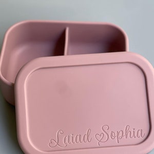 May include: A pink silicone lunch box with a divided interior and a lid that has the name "Laiad Sophia" engraved on it.