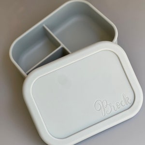 May include: A light gray silicone lunch box with three compartments. The lid has the name "Brock" embossed on it.