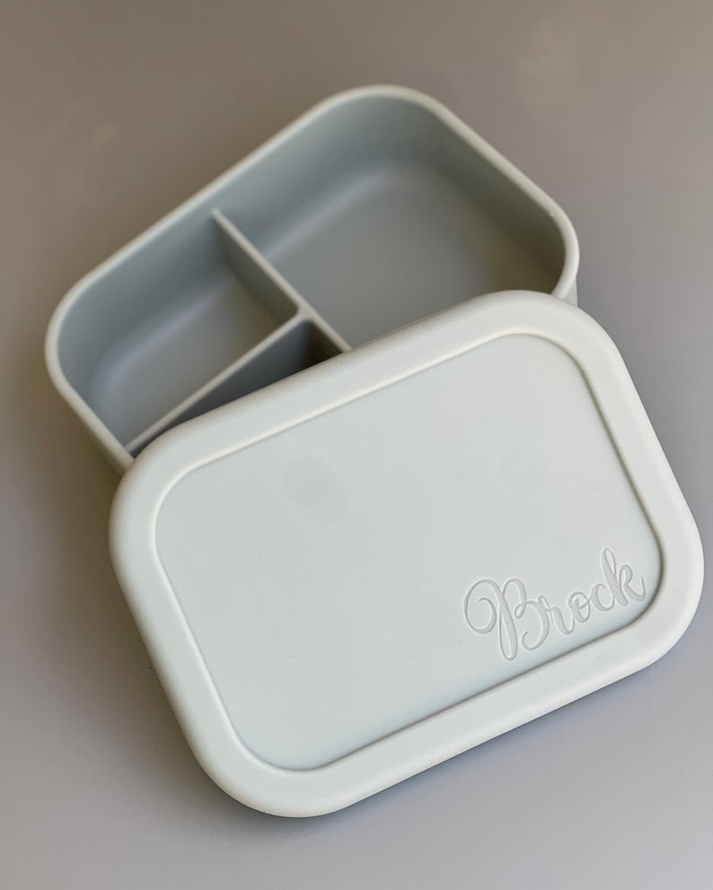 May include: A light gray silicone lunch box with three compartments. The lid has the name "Brock" embossed on it.