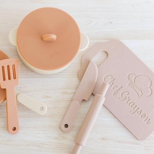 May include: A set of brown and beige silicone play kitchen tools for kids, including a pot with lid, spatula, rolling pin, knife, and chopping board. The chopping board has the text "Chef Grayson" on it.