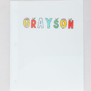 May include: A white binder with the name "GRAYSON" in colorful, apple-themed letters. The letters are outlined in black and filled with yellow, red, and blue. The binder has three rings.