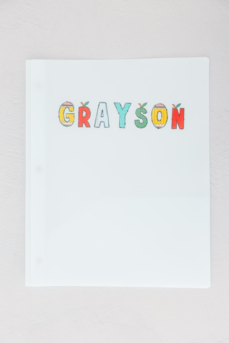 May include: A white binder with the name "GRAYSON" in colorful, apple-themed letters. The letters are outlined in black and filled with yellow, red, and blue. The binder has three rings.