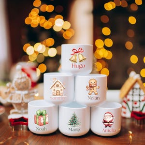 May include: White ceramic mugs with Christmas-themed illustrations and names. Designs include bells, a gingerbread house, a gingerbread man, a wrapped gift, a Christmas tree, and Santa Claus. Names on the mugs are Hugo, Sammy, Nolan, Noah, Marie, and Amelia.