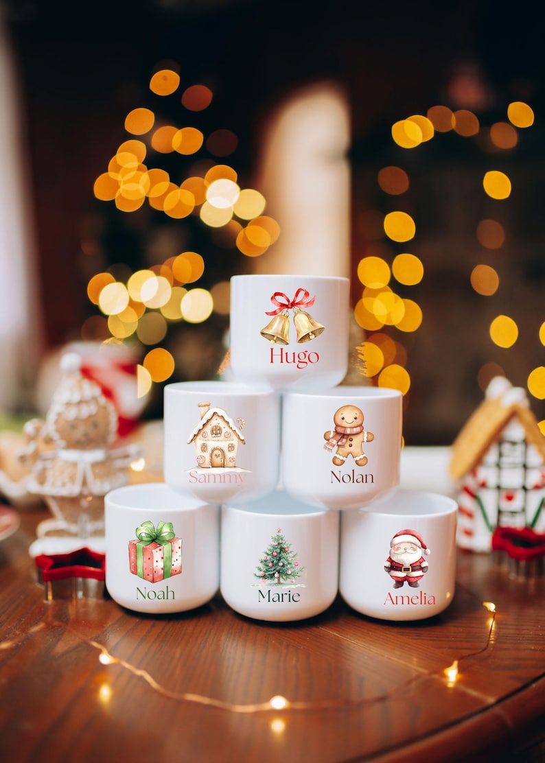 May include: White ceramic mugs with Christmas-themed illustrations and names. Designs include bells, a gingerbread house, a gingerbread man, a wrapped gift, a Christmas tree, and Santa Claus. Names on the mugs are Hugo, Sammy, Nolan, Noah, Marie, and Amelia.