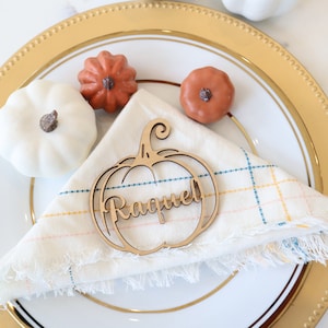 Puede incluir: Una tarjeta de lugar de madera en forma de calabaza con el nombre "Raquel" escrito en ella. La tarjeta de lugar está sobre una servilleta blanca con un patrón de cuadros. La servilleta está sobre un plato blanco con un borde dorado. Hay tres pequeñas calabazas en el plato.