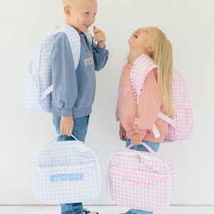 Puede incluir: Dos niños con mochilas y loncheras a juego. Las bolsas y mochilas son de cuadros vichy azul claro y blanco. Una lonchera dice "Stephen" en escritura azul, la otra "Kingsleigh" en escritura rosa.