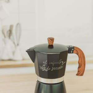 May include: Black stovetop espresso maker with a wooden handle and lid knob. The text "el cafecito de Jennie" is etched on the side. The coffee pot sits on a light-colored wooden surface.