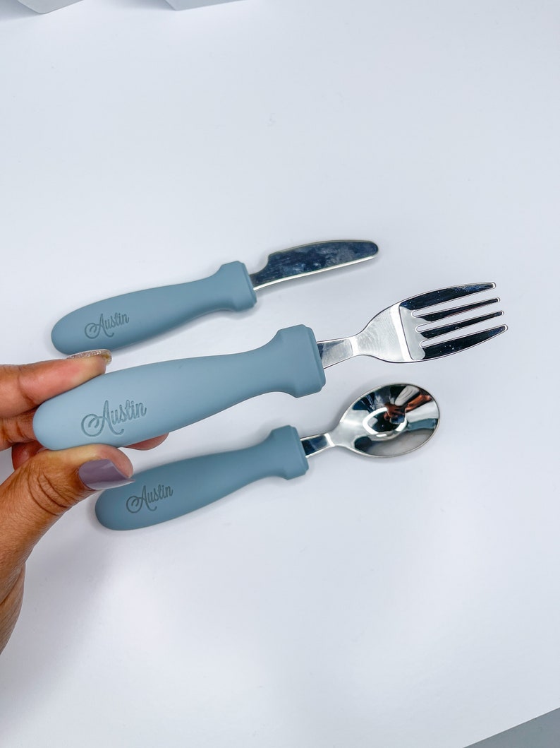 May include: A set of three blue baby utensils with the name "Austin" engraved on the handles. The set includes a fork, knife, and spoon.