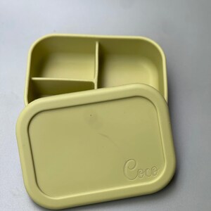 May include: A light green silicone lunch box with three compartments and a lid. The lid has the word "Coco" embossed on it.