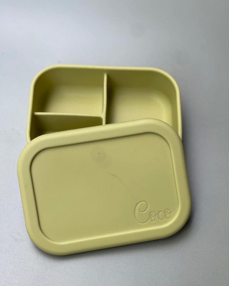 May include: A light green silicone lunch box with three compartments and a lid. The lid has the word "Coco" embossed on it.