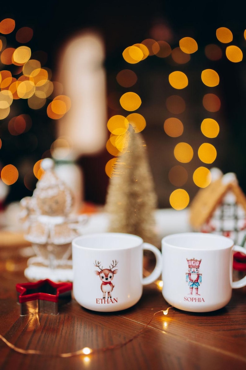 May include: Two white ceramic mugs with holiday designs. One mug features a reindeer with the name "ETHAN", the other a nutcracker with the name "SOPHIA". Festive Christmas decor includes a star cookie cutter and a small gold tree.