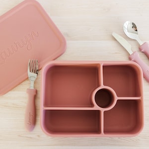 Puede incluir: Una fiambrera de color rosa polvoriento con tapa, tenedor, cuchillo y cuchara a juego. La tapa y los utensilios están grabados con el nombre "Lauryn". La fiambrera tiene cuatro compartimentos y un pozo central circular.