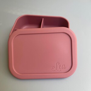 May include: A pink silicone lunch box with a divided interior and a lid that has the name "Lea" embossed on it.