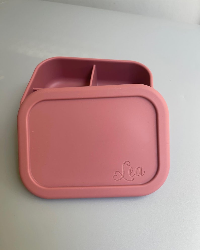 May include: A pink silicone lunch box with a divided interior and a lid that has the name "Lea" embossed on it.