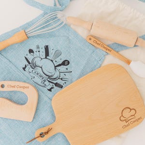 May include: A light blue apron, wooden cutting board, rolling pin, spatula, and other cooking tools. The apron has a chef hat graphic and the text "Chef Cooper". The cutting board has a chef hat graphic and the text "Chef Cooper".